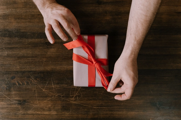 Manos atando un lazo rojo sobre un regalo envuelto en papel claro.